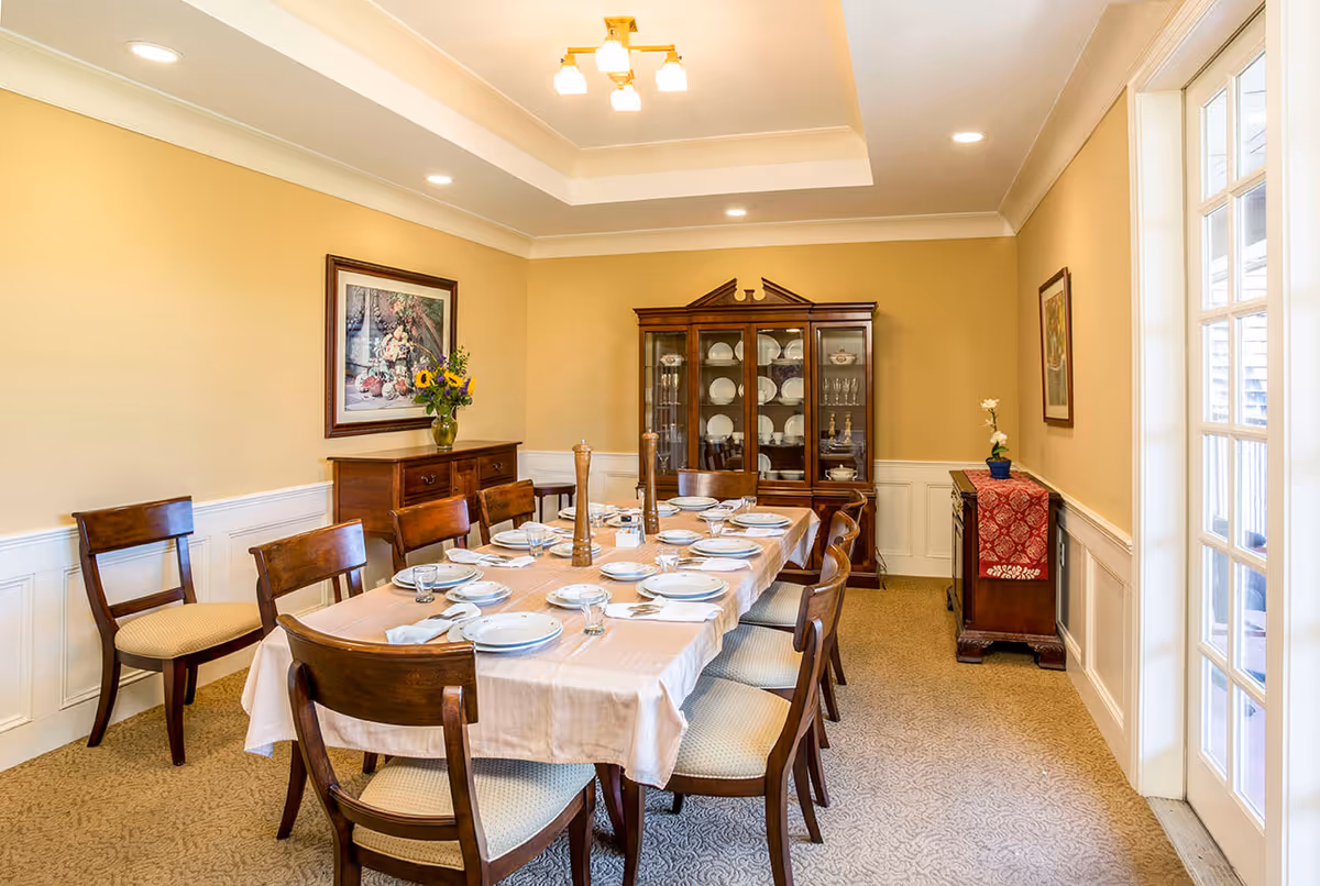 A formal dining room with a long table set for eight people with white plates, glasses, and napkins. The room features wooden chairs with cushioned seats, a wooden china cabinet displaying dishes, two sideboards with decorative items, beige walls with white wainscoting, and a ceiling light fixture. There is a framed painting on the wall and a glass door on the right side letting in natural light.