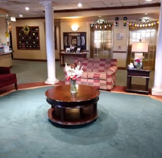 A cozy interior common area with a round wooden coffee table holding a vase of flowers in the center on a green carpet. Behind the table is a checkered sofa, a side table with a lamp and flowers, and two white columns framing the space. The background shows a bulletin board, wall decorations, and double doors with glass panels.