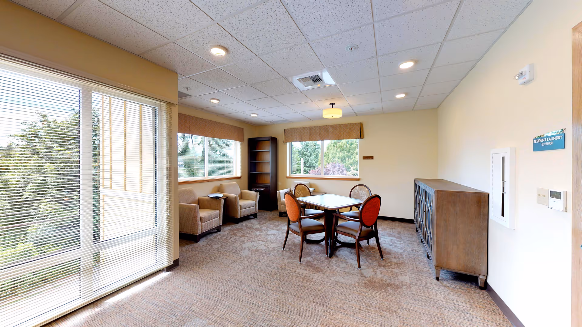 A bright room with large windows showing greenery outside. Inside, there is a square table with four chairs around it, two armchairs near the windows, a tall bookshelf, and a wooden cabinet against the wall. The room has beige walls, a carpeted floor, and ceiling lights.