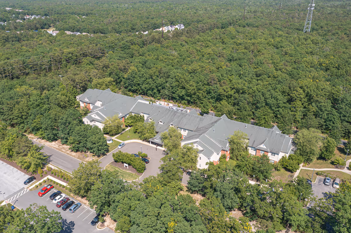 Aerial view of Woodview Estates, a large senior living facility surrounded by dense green trees. The building has a gray roof and light-colored walls with some red brick accents. There is a circular driveway with parked cars in front of the entrance, and additional parking spaces are visible nearby. The facility is located in a wooded area with a road leading to it.