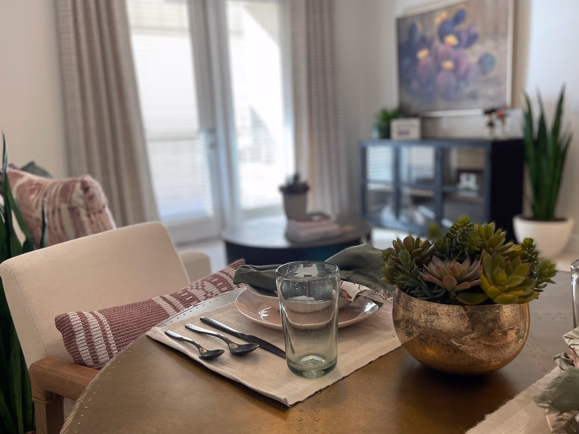 A close-up view of a dining table set with a plate, glass, cutlery, and a cloth napkin. A decorative bowl with green succulent plants is also on the table. In the background, there is a living area with a cabinet, a framed floral painting, potted plants, and large windows with curtains letting in natural light.