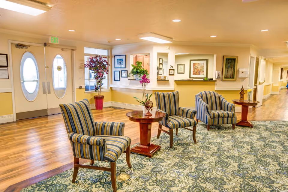A bright and welcoming seating area in a senior living facility with three striped armchairs arranged around two wooden tables. The space features a patterned carpet, wood flooring, framed artwork on the walls, and potted plants near a set of double doors with oval windows. A hallway extends to the right.