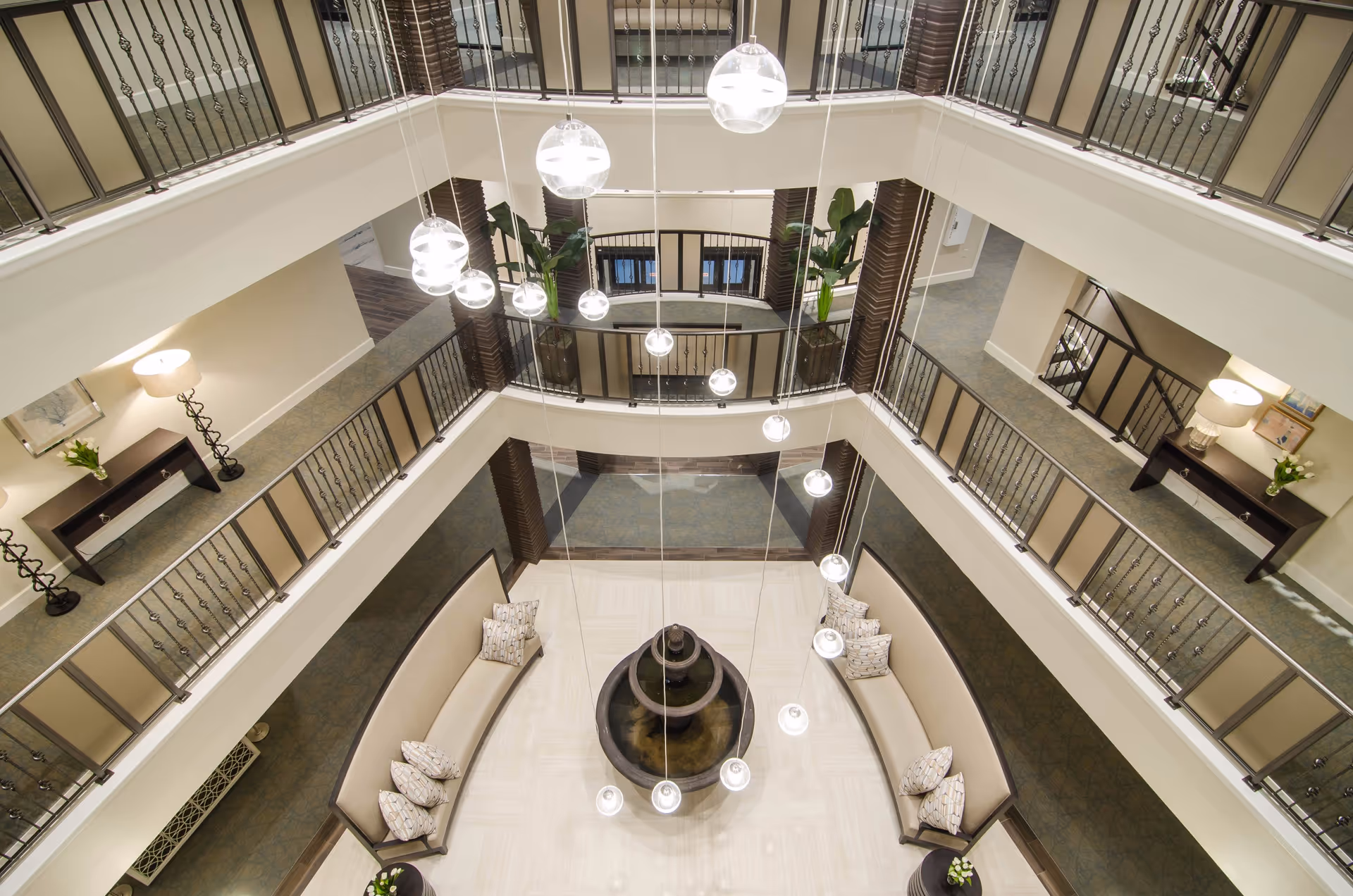 View from above of a multi-level indoor common area with curved beige sofas and patterned pillows surrounding a central water fountain. Modern hanging globe lights descend from the ceiling. The area is bordered by railings and decorated with plants and lamps on side tables.