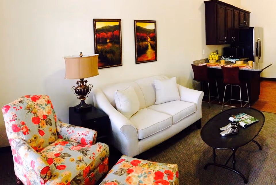 A cozy living room area featuring a white sofa with two white pillows, a floral patterned armchair with a matching ottoman, a dark wooden side table with an ornate lamp, and two framed landscape paintings on the wall. In the background, there is a kitchen area with dark wood cabinets, a granite countertop with three bar stools, a microwave, and a refrigerator. A black oval coffee table in front of the sofa holds a magazine and a pair of silver shoes.