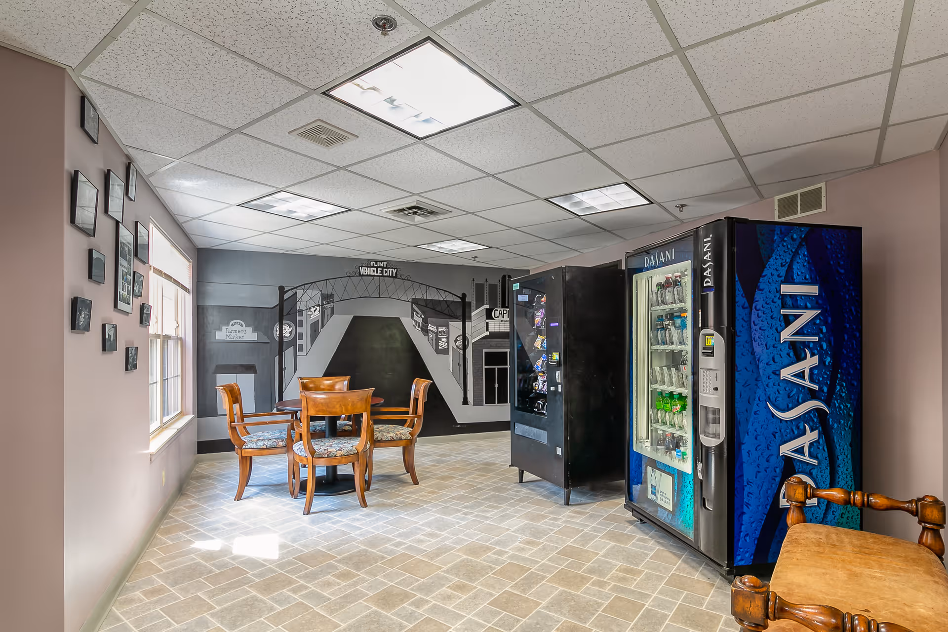 A small common area with a round wooden table and four chairs with floral cushions. There are two vending machines, one for snacks and one for Dasani bottled water, against the wall. The wall behind the table features a mural depicting a street scene with a sign that reads 'Flint Vehicle City.' Several framed pictures are hung on the adjacent wall near a window with blinds. The floor is tiled with a light-colored pattern, and the ceiling has recessed fluorescent lighting.