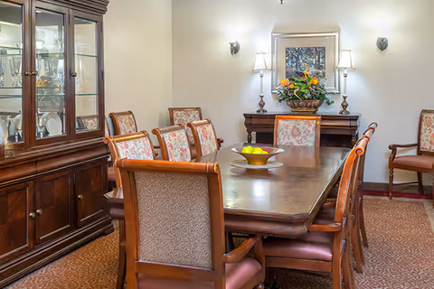 A formal dining room with a long wooden table surrounded by upholstered chairs. A wooden cabinet with glass doors displaying glassware is on the left. At the far end of the room, there is a sideboard with a floral arrangement and a framed picture above it. The room is softly lit by wall sconces and table lamps.