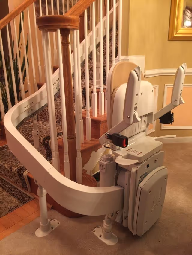 A stairlift installed on a curved staircase with wooden handrails and white balusters in a warmly lit interior space with beige walls and carpeted floor.
