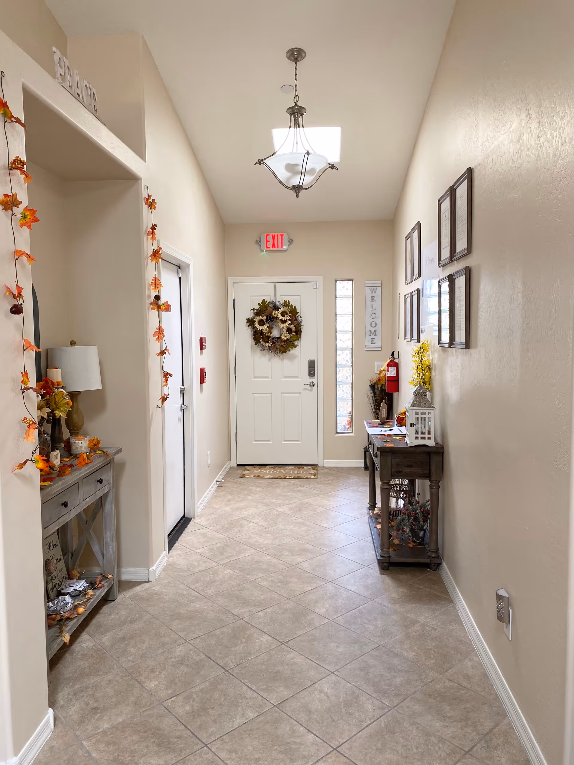 A well-lit hallway with beige walls and tiled floor leading to a white door decorated with a floral wreath. On the left side, there is a small table with a lamp and autumn-themed decorations, and on the right side, a narrow table with framed pictures on the wall above it and additional decorative items. A ceiling light fixture hangs in the center, and an exit sign is visible above the door.