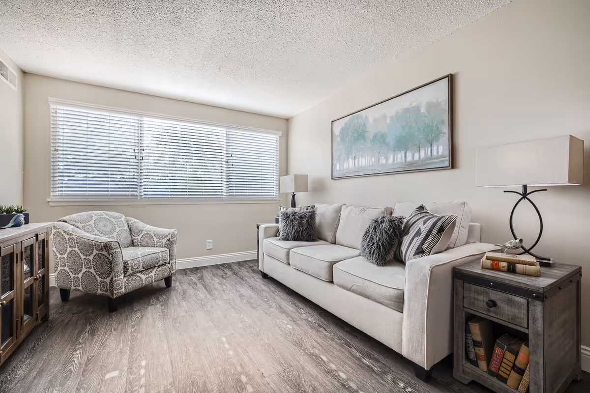 Well-lit living room featuring a beige sofa with decorative pillows, a patterned armchair, side tables with lamps, and a wide window with blinds.