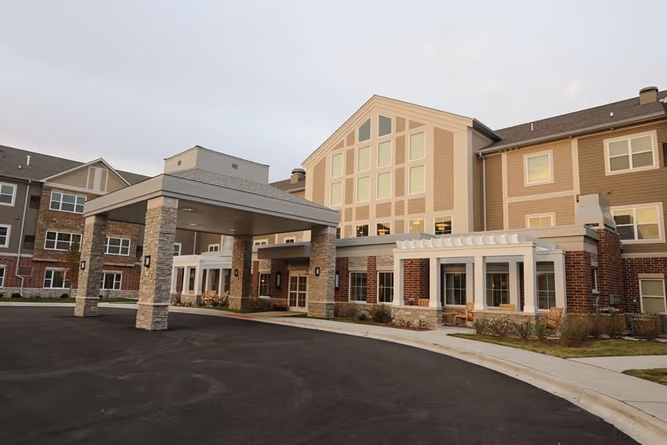 Exterior view of Clarendale of Mokena, a multi-story senior living facility with a covered entrance supported by stone pillars, large windows, and outdoor seating areas with benches along the front.