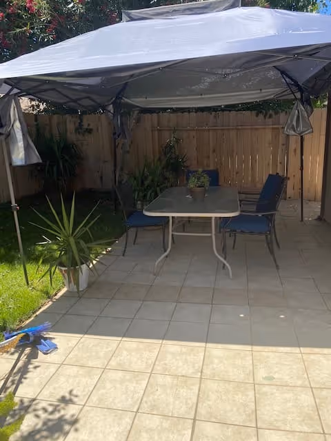 Tiled backyard patio with a canopy-covered table, chairs, potted plants, and a wooden fence.