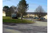 Exterior view of Clinton Manor Apartments showing a parking lot, a grassy area with trees, and a two-story building in the background under a clear sky.
