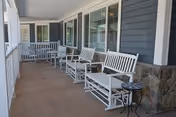 A covered porch area with white wooden benches and rocking chairs arranged along the side of a building with gray siding and large windows. A small round black metal table is placed near one of the benches. The porch floor is made of wood and there is a stone column at the corner.
