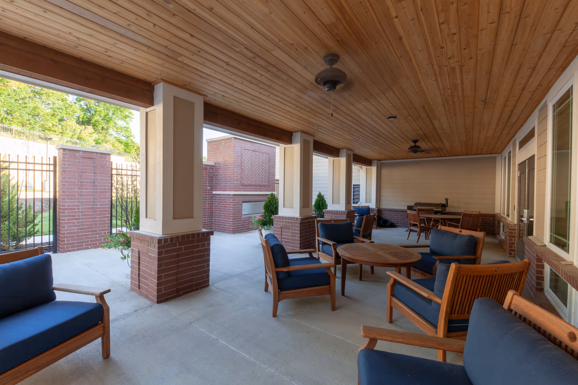 Covered outdoor patio area with wooden ceiling and ceiling fans, furnished with wooden chairs and sofas with blue cushions, round wooden tables, and brick pillars. The patio overlooks a fenced garden area with greenery.