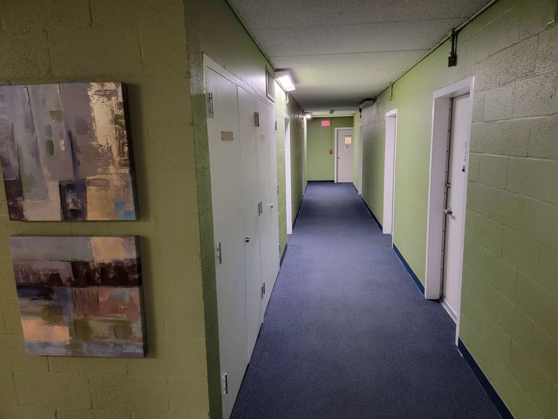 Carpeted interior hallway with green painted cinderblock walls, multiple doors along the corridor and framed artwork on the left wall.