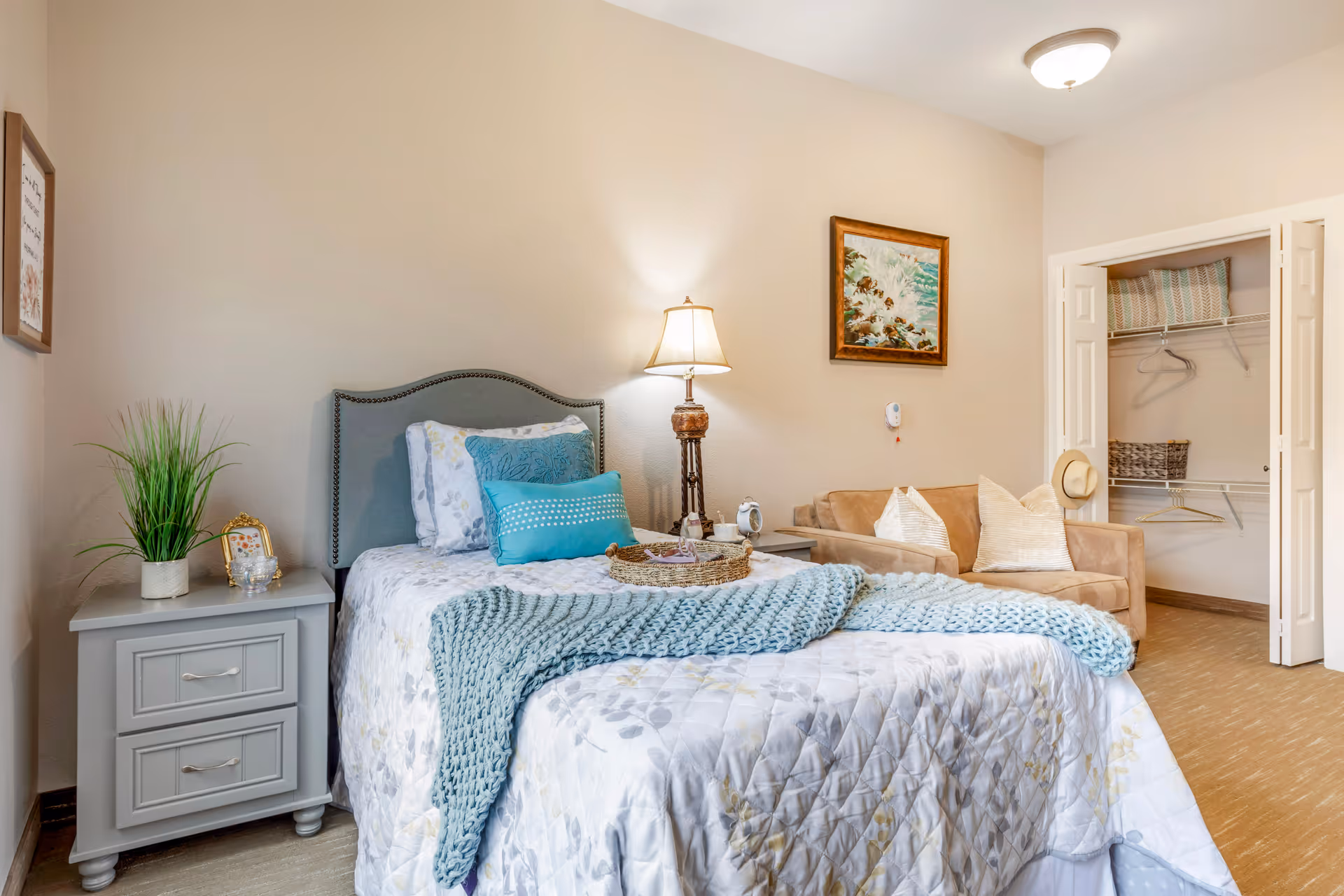 A cozy bedroom in a senior living facility featuring a single bed with a gray upholstered headboard, white and blue bedding, and a light blue knitted throw blanket. Next to the bed is a gray nightstand with a potted plant, a small framed picture, and a glass candle holder. A tall lamp with a beige shade is positioned between the bed and a beige loveseat with two cream-colored pillows. On the wall above the loveseat is a framed painting of ocean waves crashing on rocks. An open closet with white bi-fold doors reveals shelves with pillows, baskets, and hangers. The room has beige walls and carpeted flooring.
