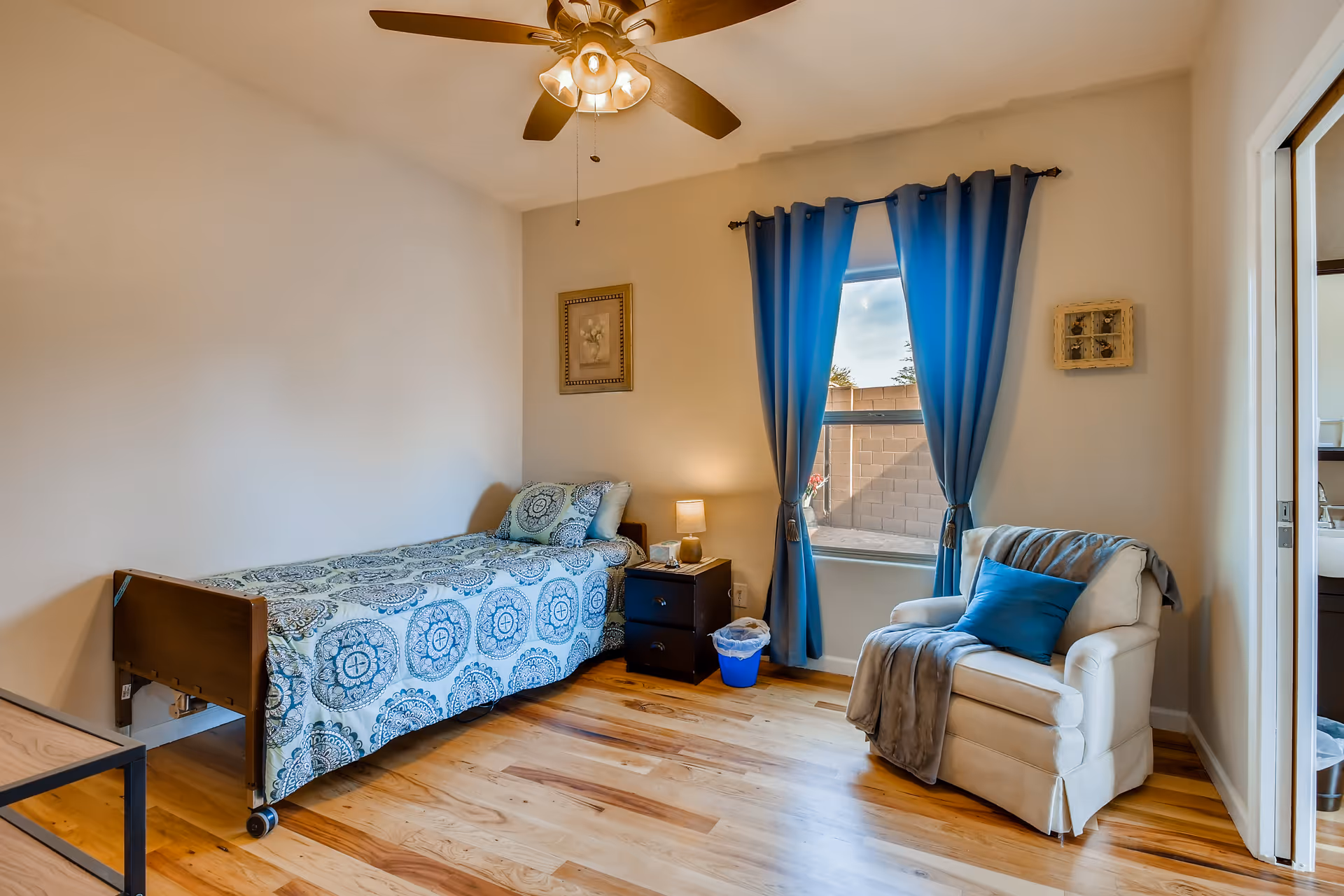 A cozy bedroom with a single bed covered in a blue patterned bedspread, a nightstand with a lamp, a white armchair with a blue pillow and gray throw, blue curtains on the window, and a ceiling fan with lights. The room has wooden flooring and light-colored walls.