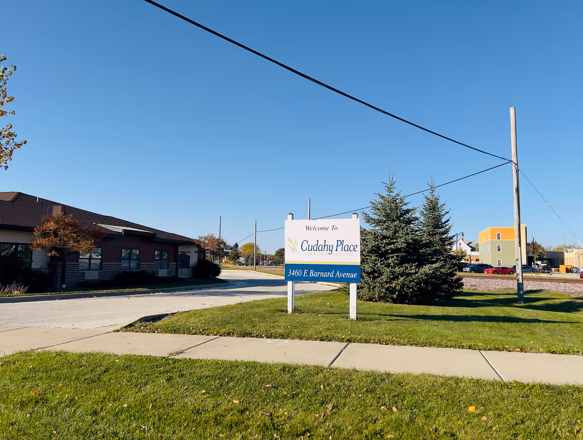 Outdoor view of Cudahy Place Senior Living facility with a sign displaying the name and address 3460 E Barnard Avenue, a building on the left, green grass, trees, and a clear blue sky.