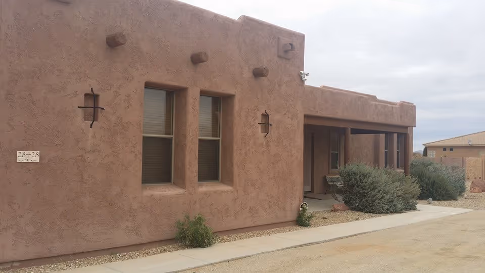 Exterior view of a southwestern-style building with stucco walls and two windows. There is a covered porch area with bushes and a concrete walkway in front. The sky is overcast.