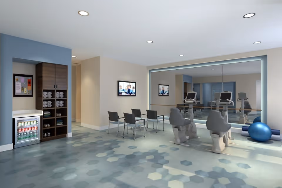 A modern fitness room with exercise bikes facing a large mirror on the wall. There are four chairs arranged in a row, a small refrigerator stocked with beverages, shelves with rolled towels, and exercise equipment including a blue exercise ball and yoga mats. The room has a hexagonal patterned floor and recessed ceiling lights.