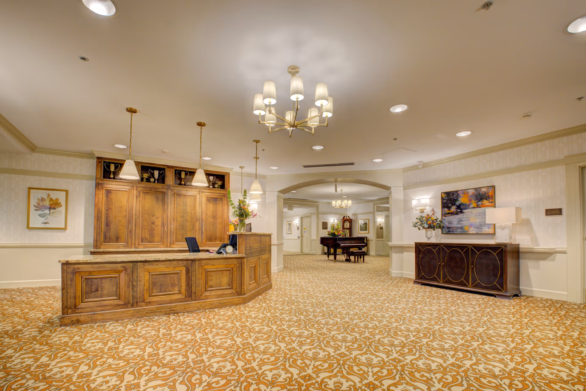 Spacious and well-lit reception area with a wooden front desk, decorative carpet, chandelier lighting, a piano in the background, and artwork on the walls.