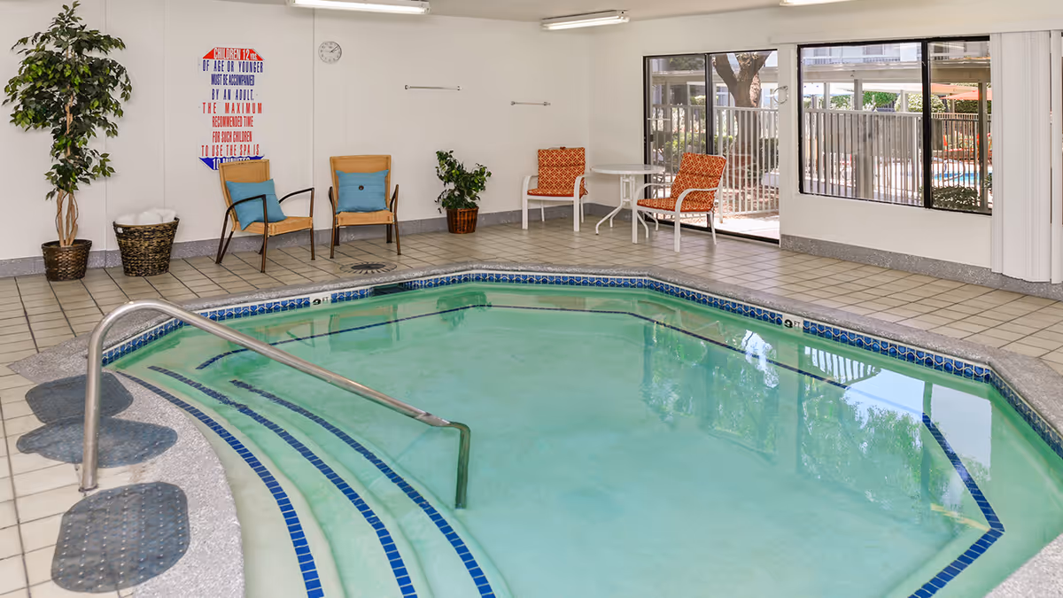 Indoor pool area with clear water, tiled floor, metal handrail, and steps leading into the pool. There are several chairs with cushions and small tables along the walls, potted plants, and large windows showing an outdoor fenced area with more seating and umbrellas.