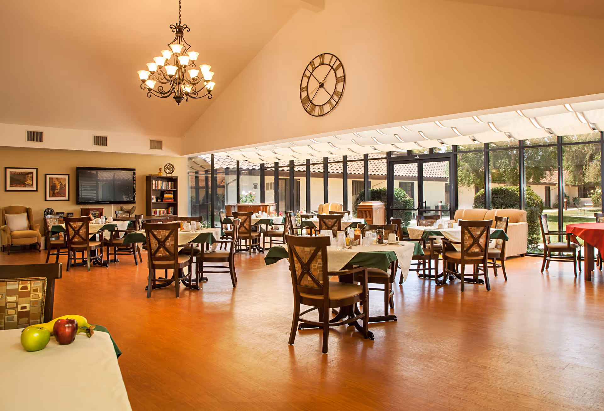 A spacious dining room with multiple tables covered with white and green tablecloths, set with cups and glasses. The room has large floor-to-ceiling windows letting in natural light and offering a view of a garden outside. There is a large decorative clock on the wall, a chandelier hanging from the ceiling, a TV mounted on the wall, and comfortable chairs arranged around the tables.