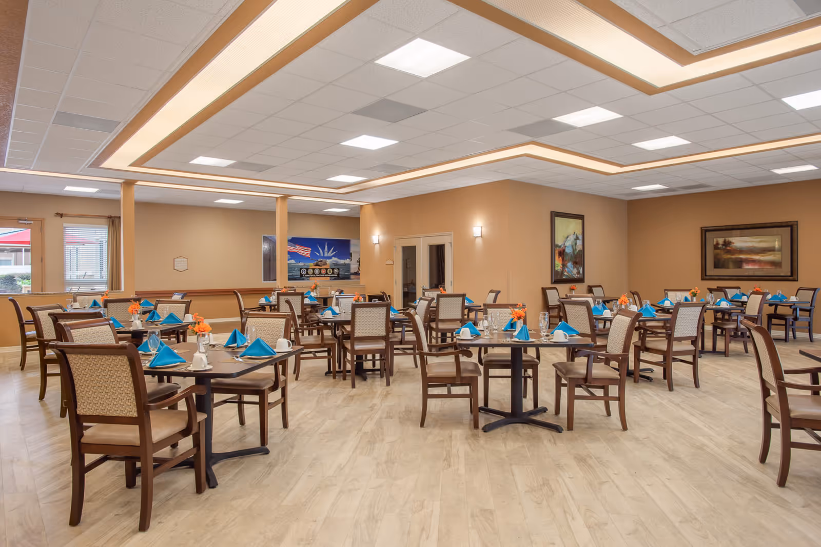 A spacious dining room with multiple tables set with blue napkins, white cups, and small orange flower centerpieces. The room has beige walls, light wood flooring, and ceiling lights with a decorative recessed lighting feature. There are framed paintings on the walls and windows with curtains on the left side.