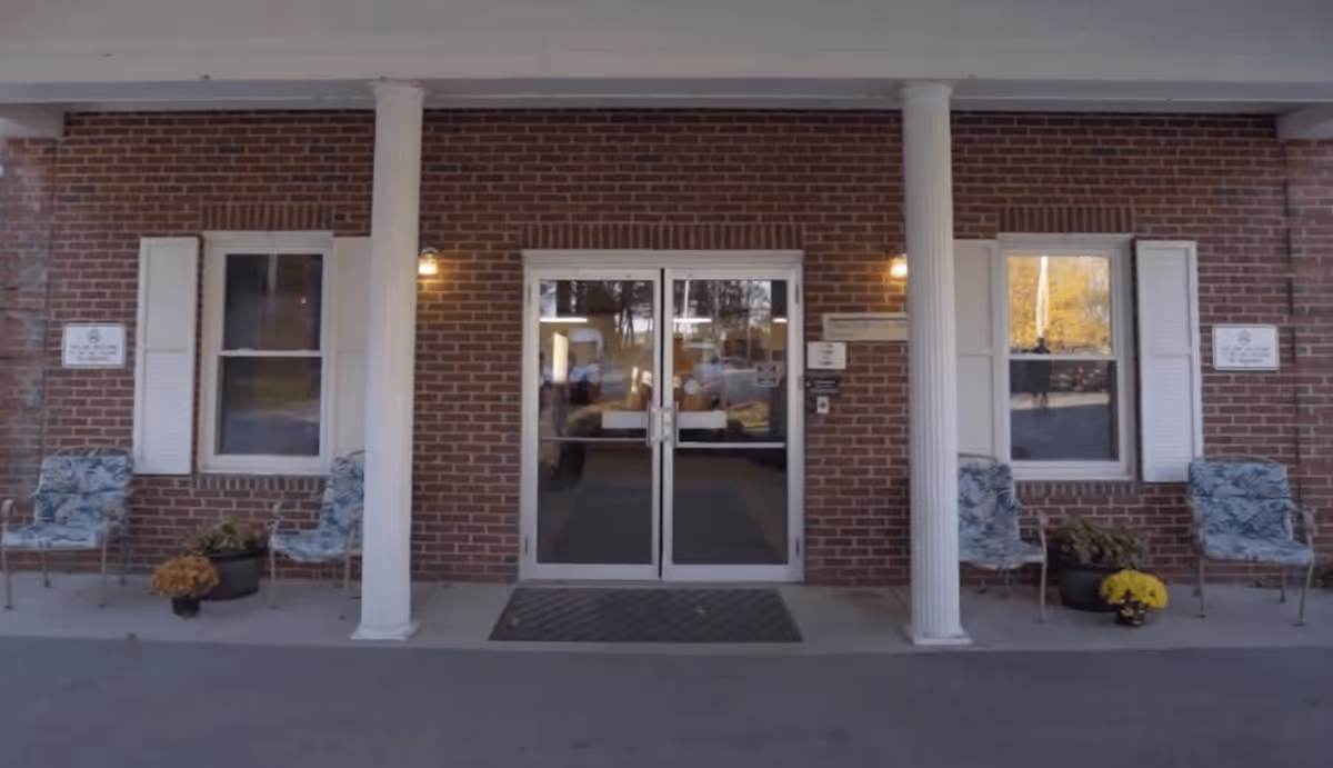 Front entrance of a brick building with two white columns flanking glass double doors. There are two windows with white shutters on either side of the doors, each accompanied by two patterned chairs and potted plants with flowers. Wall-mounted lights illuminate the entrance area.
