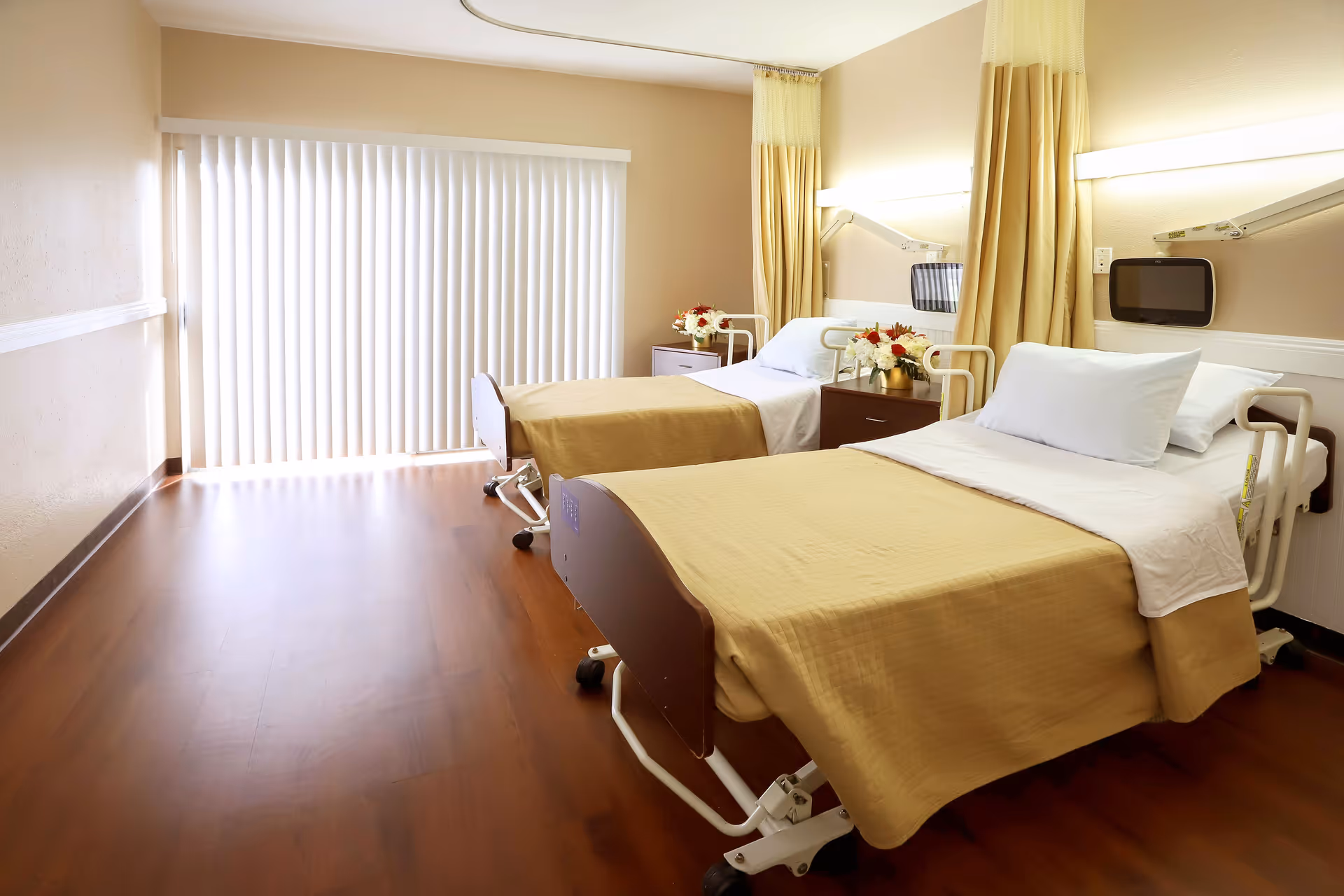 A bright and clean room with two hospital-style beds, each covered with beige blankets and white pillows. Between the beds is a small nightstand with a flower arrangement. The room has wooden flooring and a large window with vertical blinds letting in natural light. Beige curtains hang on the wall behind the beds.