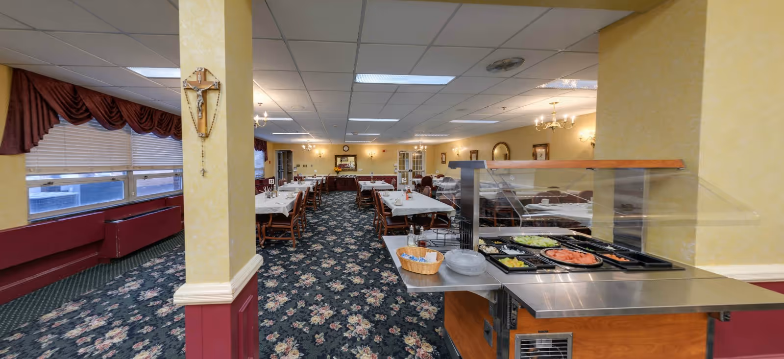 Interior view of a dining room in a senior living facility with tables covered in white tablecloths, wooden chairs, floral carpet, and a salad bar with various fresh vegetables. The room has yellow walls, chandeliers, and large windows with maroon valances.