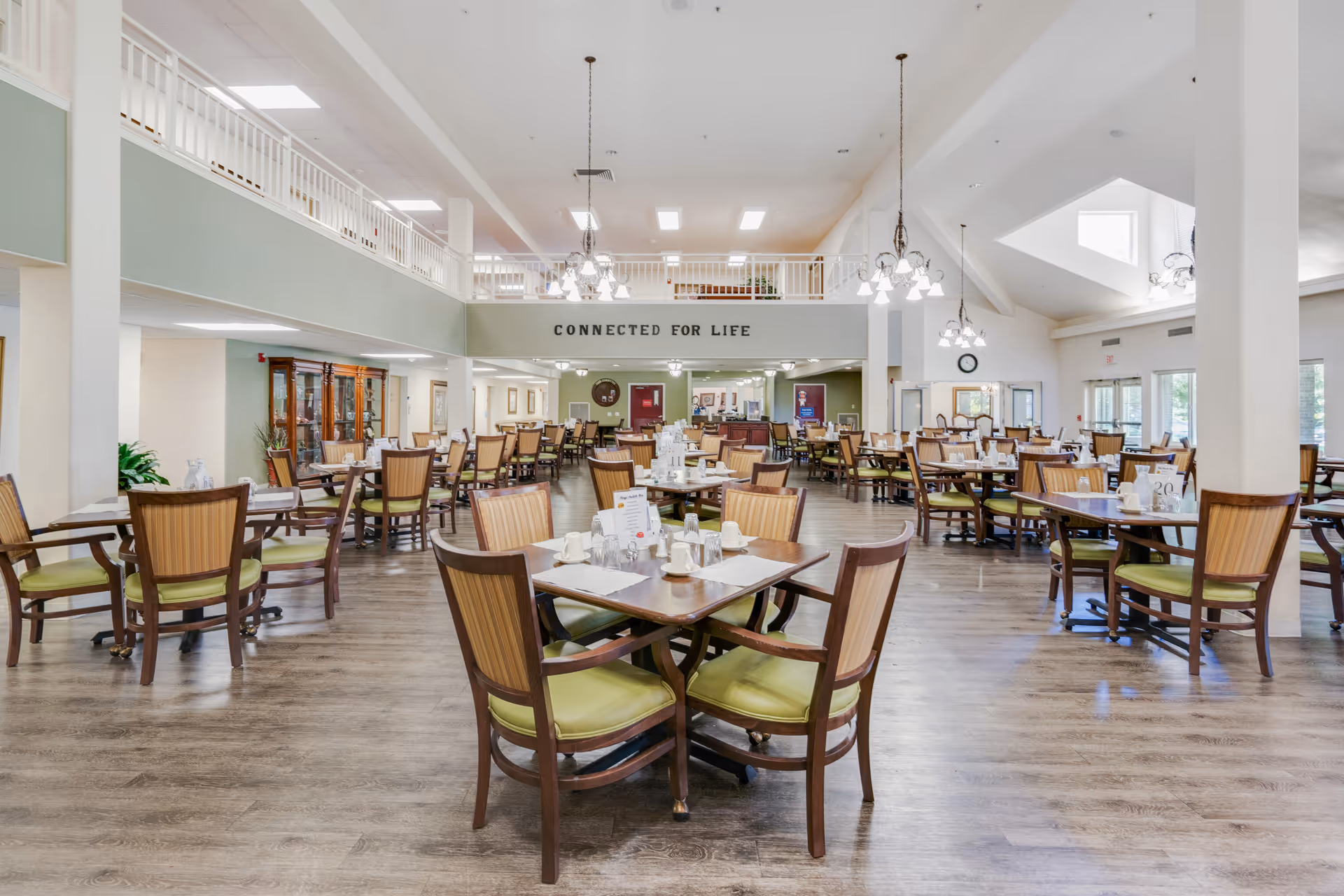 Spacious dining room with multiple wooden tables and chairs arranged neatly. The room has high ceilings with hanging light fixtures and large windows allowing natural light. A balcony railing overlooks the dining area, and a sign on the balcony reads 'CONNECTED FOR LIFE'.