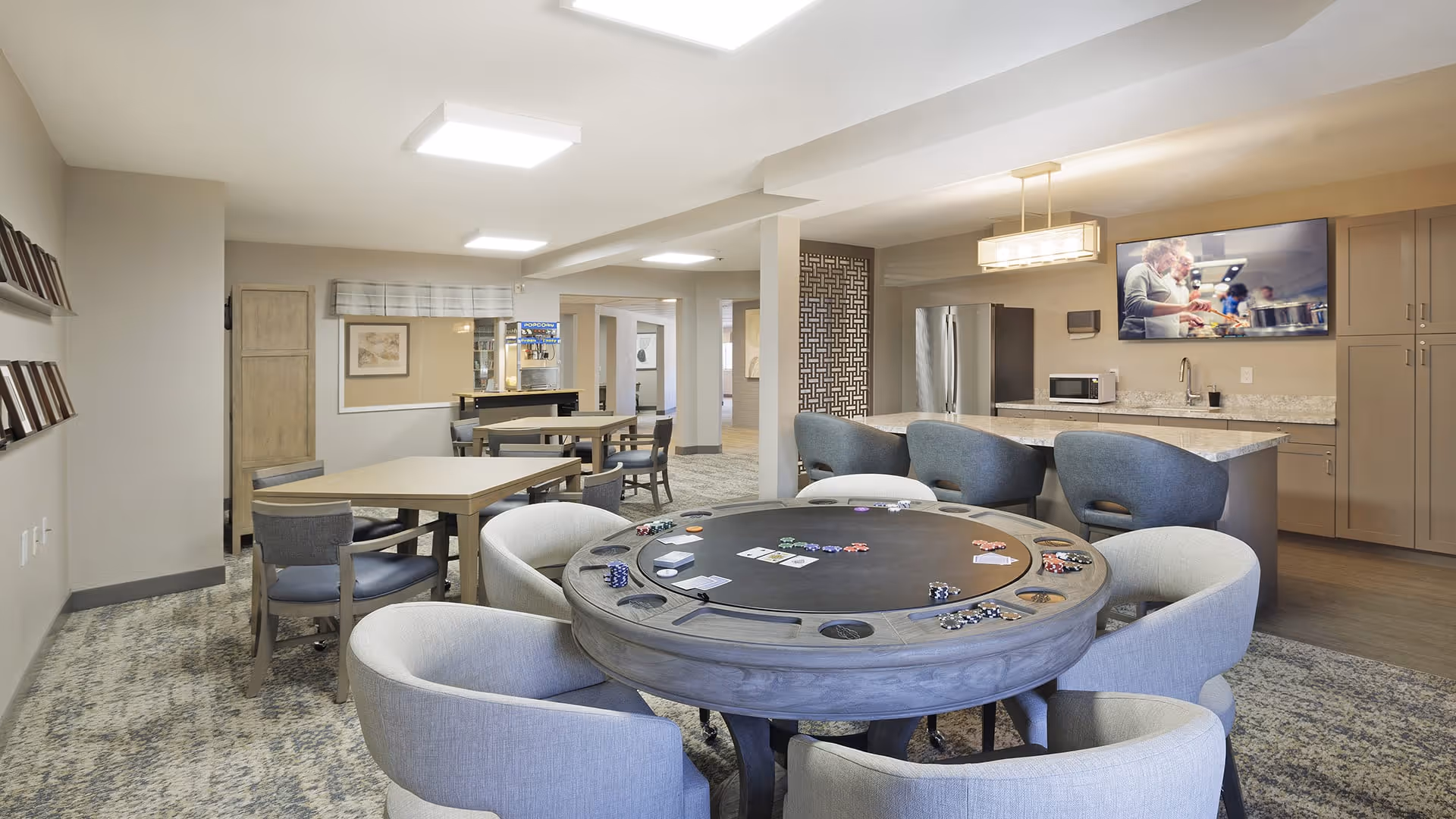 A spacious common area in a senior living facility featuring a round poker table with chips and cards, surrounded by six cushioned chairs. In the background, there are additional tables and chairs, a kitchenette with a refrigerator, microwave, and cabinets, and a wall-mounted TV displaying an image of two elderly people cooking together.