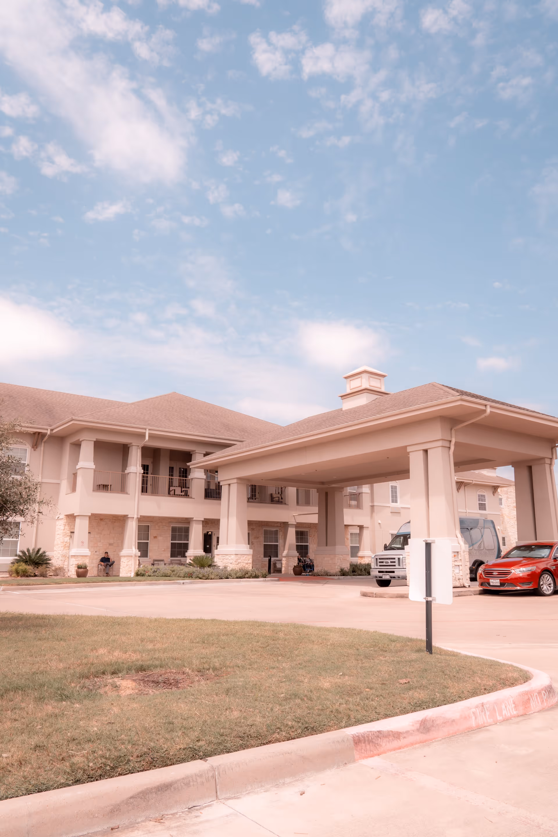 Front exterior of a two-story senior living building with a covered drop-off portico, parked vehicles, and a blue sky.