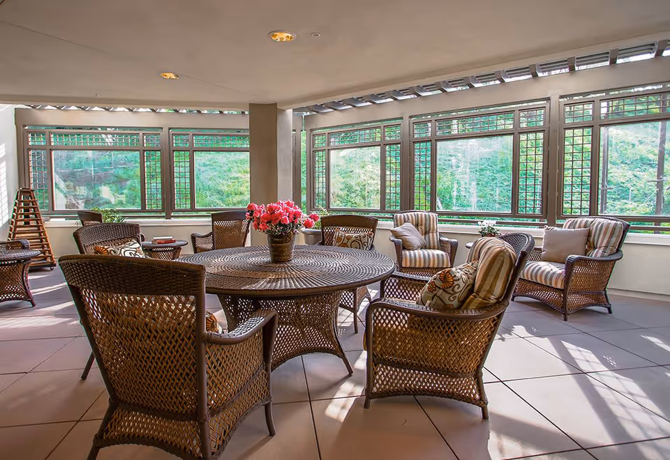 A bright indoor seating area with large windows showing greenery outside. The space features a round wicker table with a flower vase centerpiece and several wicker armchairs with patterned cushions arranged around it.