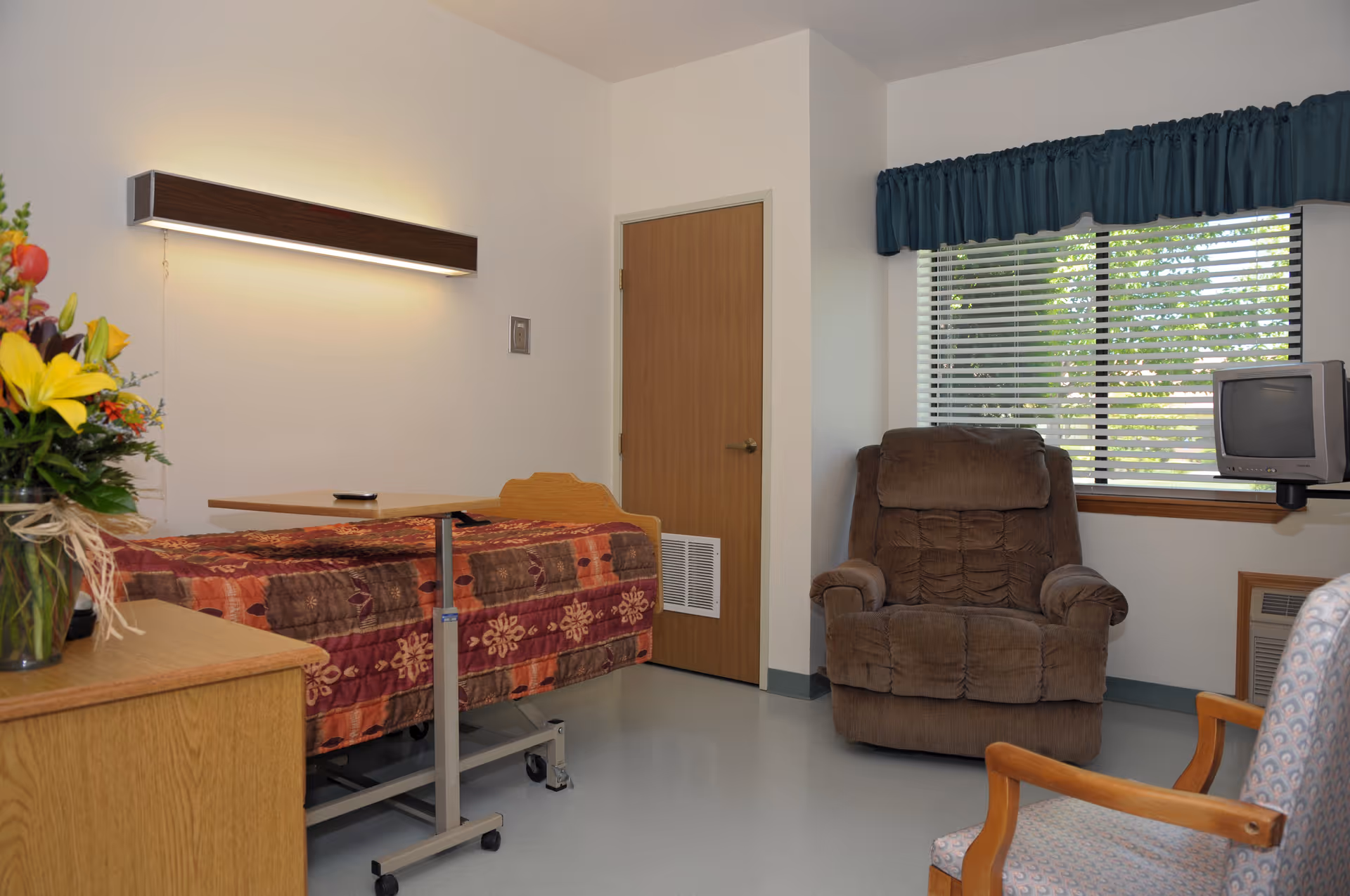 A senior living facility room with a single bed covered by a patterned quilt, a wooden overbed table, a brown recliner chair, a wooden dresser with a vase of colorful flowers, a window with blinds and a blue valance, and a small television mounted near the window.
