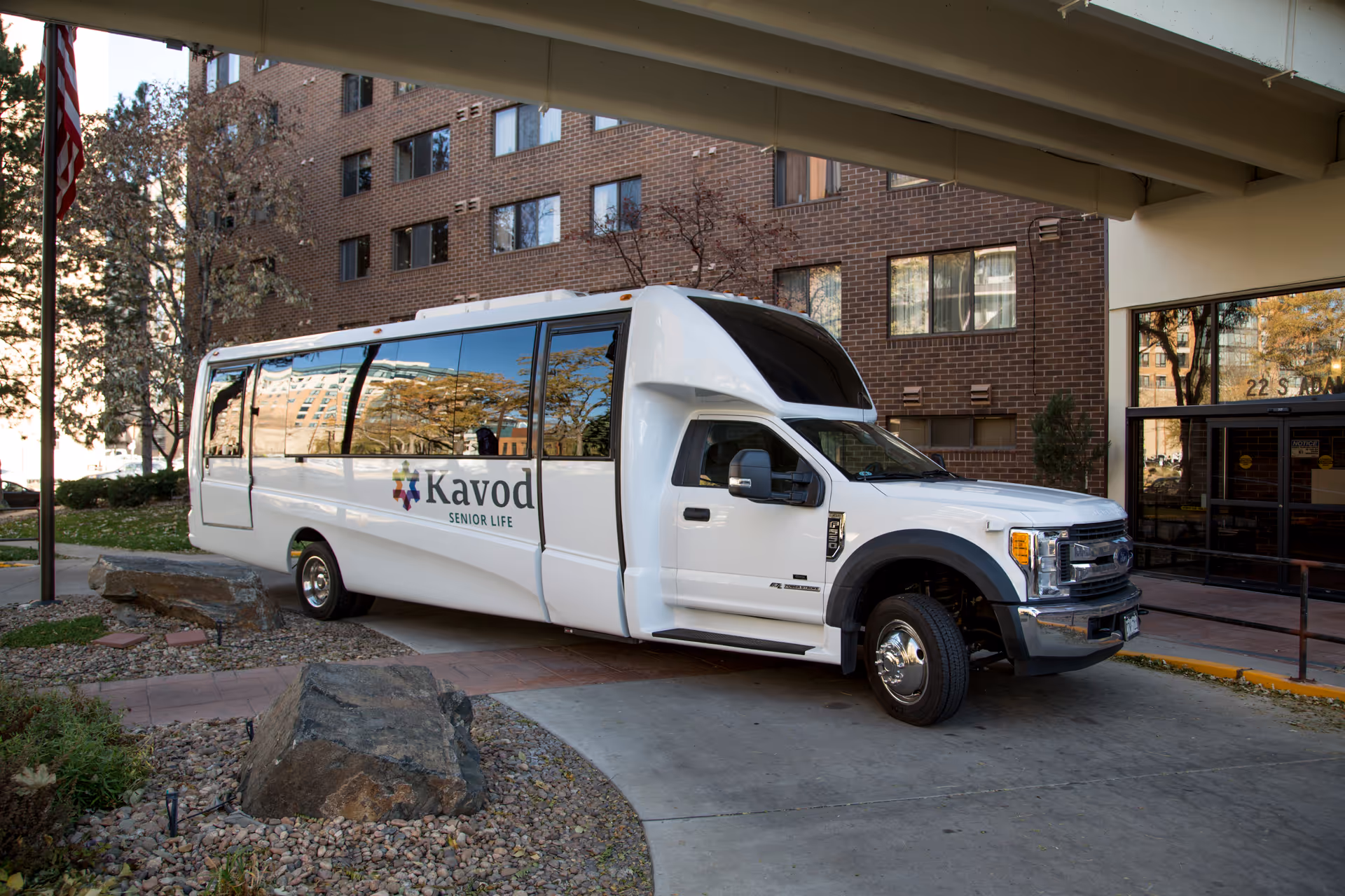White Kavod Senior Life shuttle parked under a canopy at the entrance of a brick senior living building.