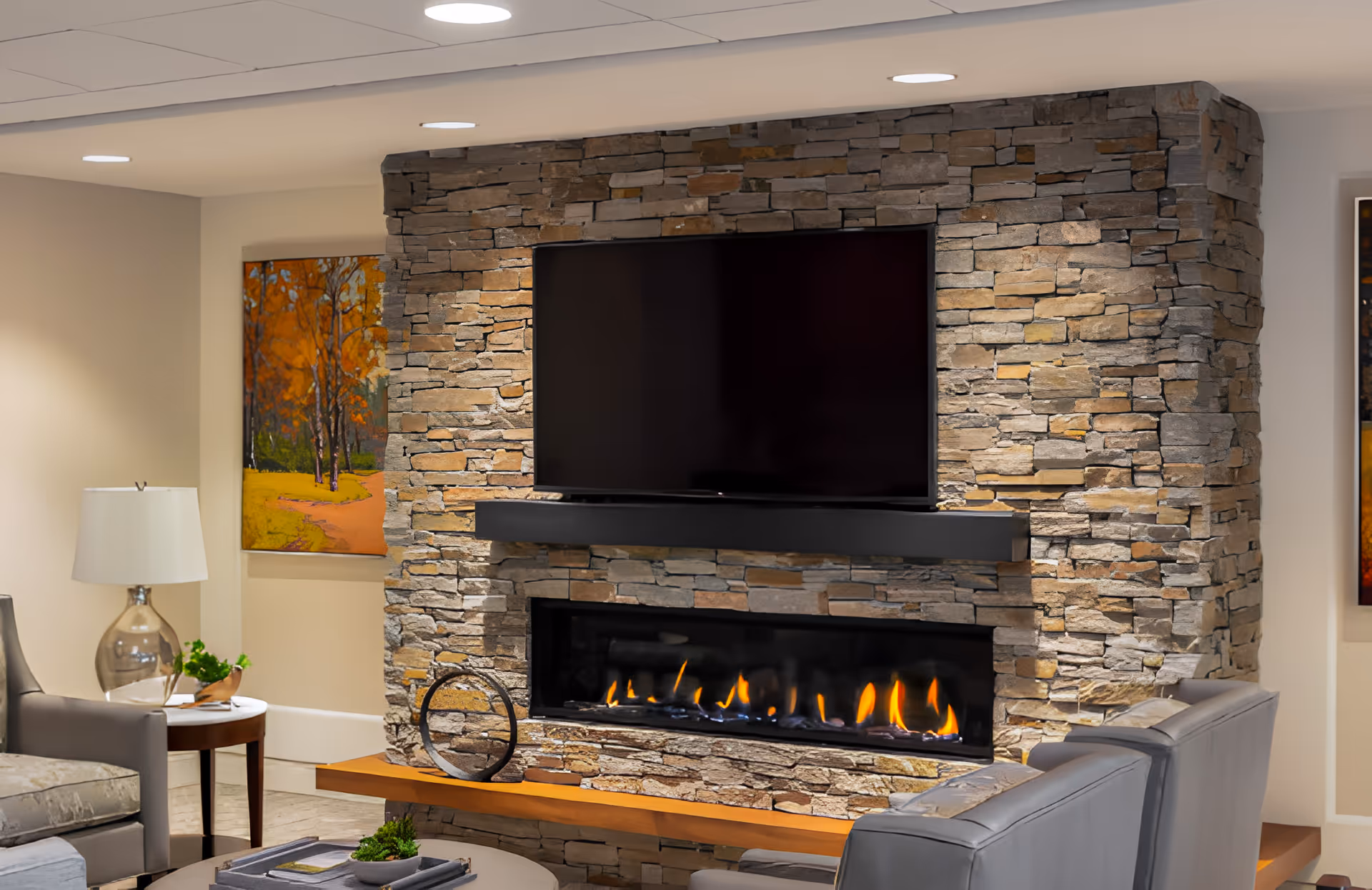 Cozy living room area featuring a stone fireplace with a modern linear gas fire and a large flat-screen TV mounted above it. The room includes comfortable gray armchairs, a round side table with a glass lamp and small plant, and colorful autumn-themed artwork on the walls.