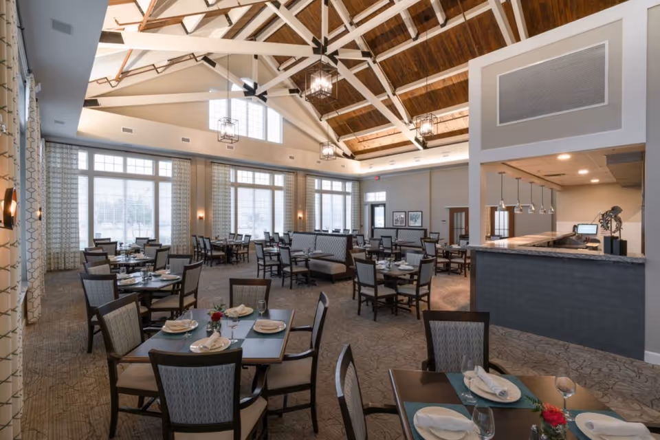 A spacious dining room with multiple tables set with plates, napkins, glasses, and silverware. The room features large windows with patterned curtains, high vaulted ceilings with exposed wooden beams, and modern hanging light fixtures. There is a serving counter area on the right side of the image.
