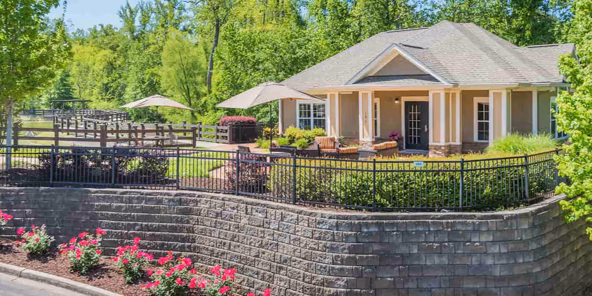 A single-story building with a covered porch surrounded by a black metal fence and landscaped greenery. There are outdoor seating areas with umbrellas and chairs on a paved patio. The scene is bright and sunny with trees and bushes in the background.