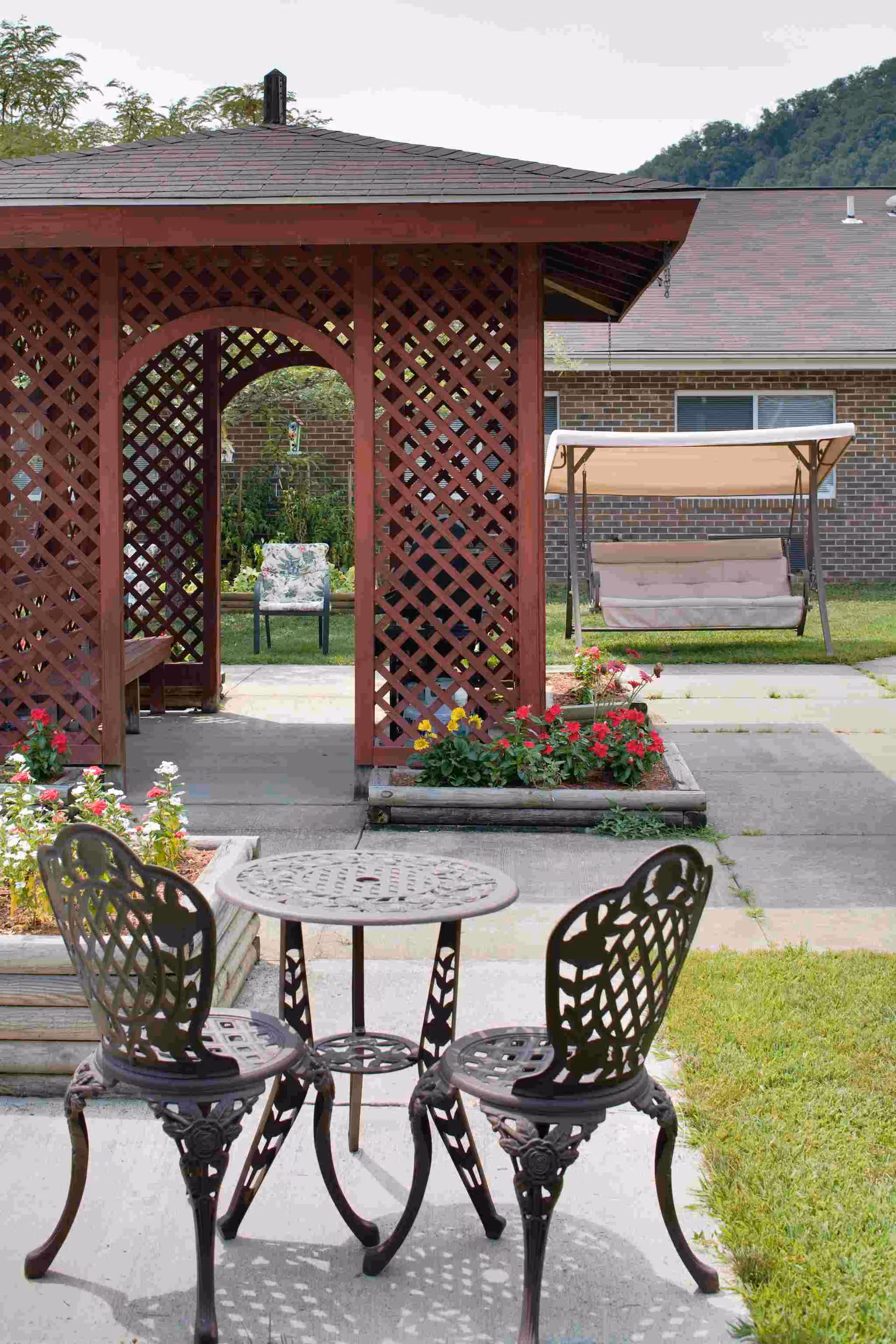 Outdoor patio area with a small round metal table and two matching chairs in the foreground. Behind them is a wooden gazebo with lattice sides and a bench inside. There are flower beds with colorful flowers around the gazebo. In the background, there is a cushioned swing with a canopy and a brick building with windows. Trees and hills are visible in the distance.