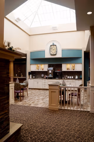 Bright interior common area with a skylight over a coffee station and cabinets, a large clock, and small dining tables and chairs.
