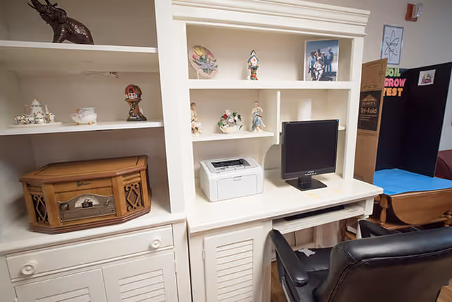A white built-in desk and shelving unit with various decorative items including figurines and a photo frame. On the desk are a computer monitor and a printer. A black office chair is positioned in front of the desk. To the right, there is a cardboard box and a bulletin board with colorful text.