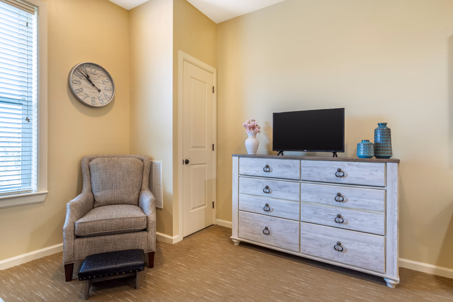 Armchair with ottoman beside a window faces a whitewashed dresser topped with a TV and decorative vases in a neutral-toned room.