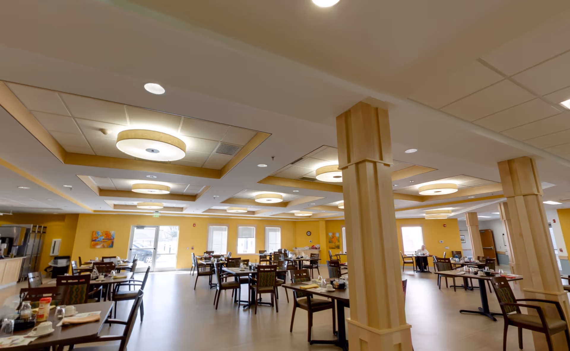 Spacious senior living dining room with multiple tables and chairs, warm yellow walls, wooden columns, and recessed ceiling lights.