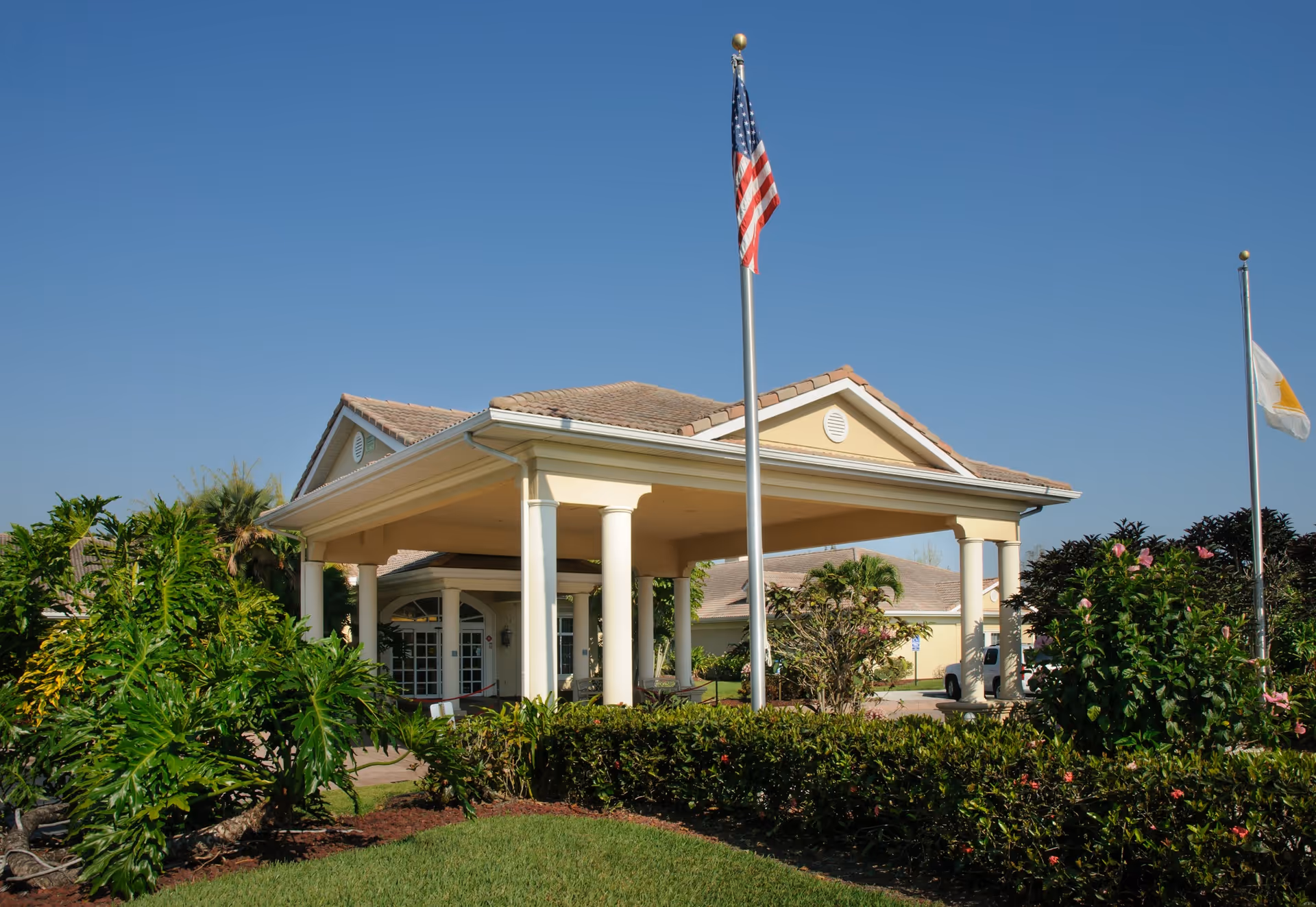 Front exterior view of a single-story building with a covered entrance supported by white columns. There are two flagpoles in front, one with an American flag and the other with a white flag. The building is surrounded by well-maintained greenery and bushes under a clear blue sky.