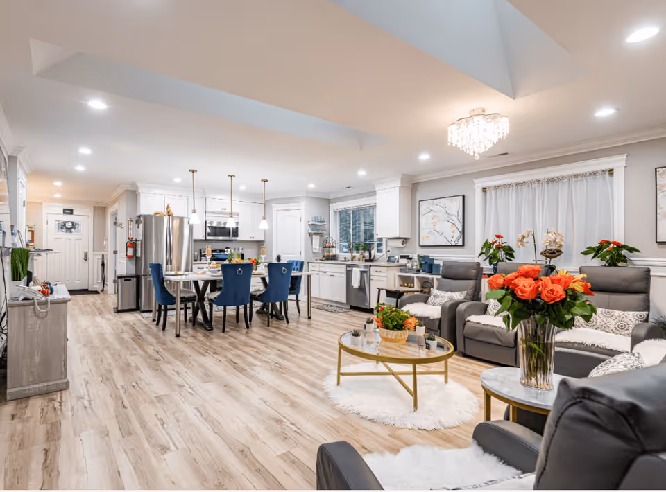 A spacious and modern open-concept living area in an adult family home featuring a kitchen with white cabinets, stainless steel appliances, and a dining table with blue chairs. The living room area has gray recliner chairs, a glass coffee table with a vase of orange roses, and decorative plants and artwork on the walls. The flooring is light wood, and the room is well-lit with recessed lighting and a chandelier.