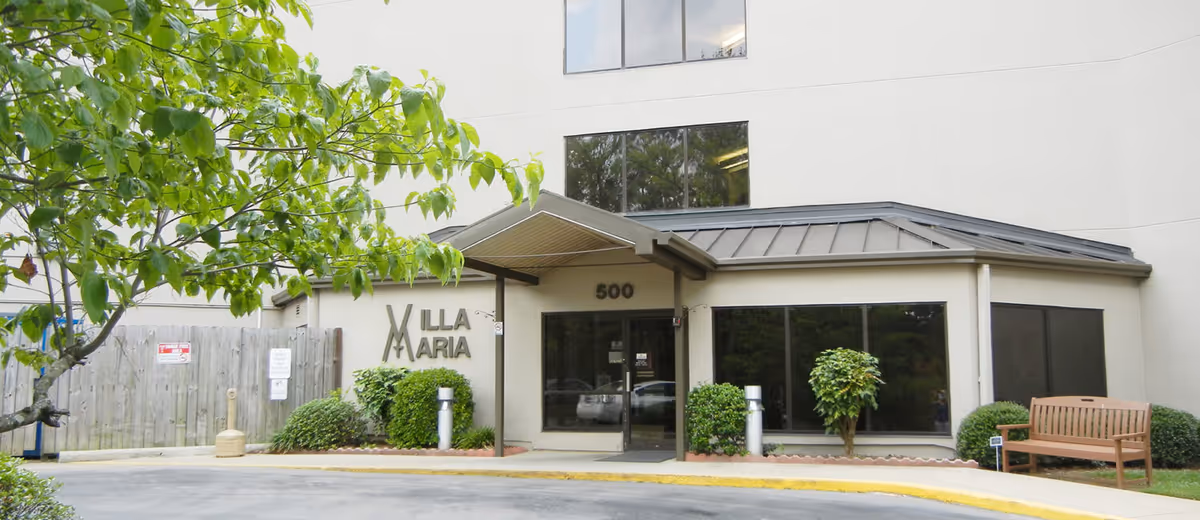 Front exterior of Villa Maria with entrance, greenery, and a wooden bench.