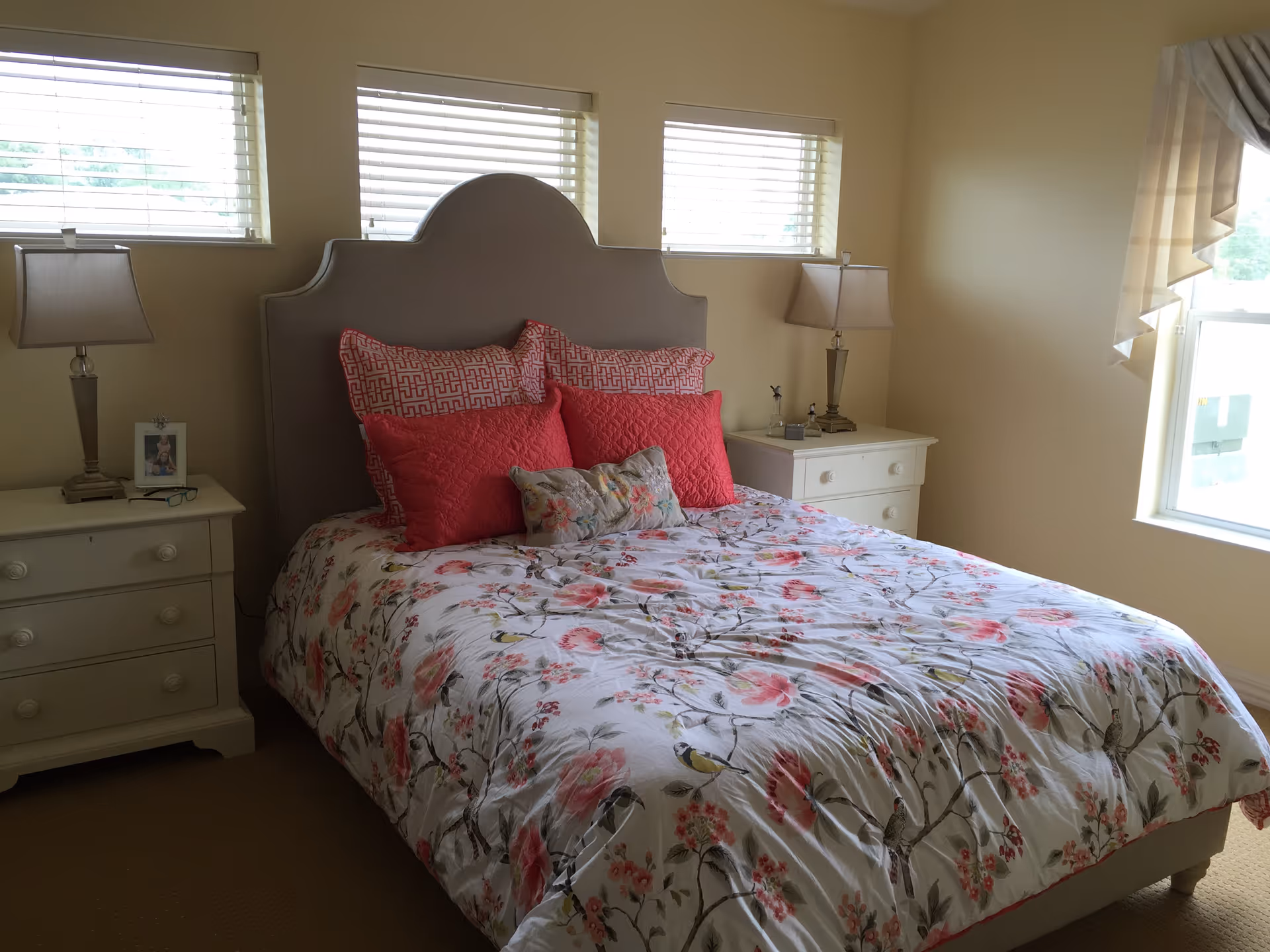 Sunlit bedroom with a floral bedspread, upholstered headboard, and matching nightstands with lamps.