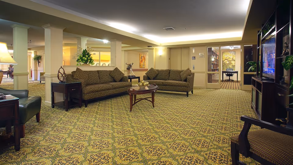 A spacious senior living community lounge area with patterned carpet, two brown patterned sofas, a wooden coffee table with decorative items, a large TV on a dark wooden entertainment center, and a well-lit ceiling. The room has beige walls with columns and a side table with a lamp and decorative sculpture. In the background, there is an office area with glass doors and a painting on the wall.